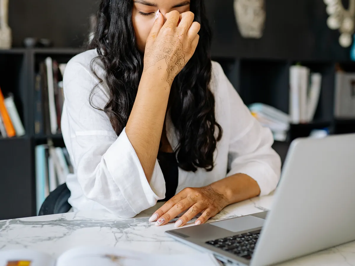 Femme devant un ordinateur portable, les yeux fermés et une main sur le visage, avec des documents sur le bureau.